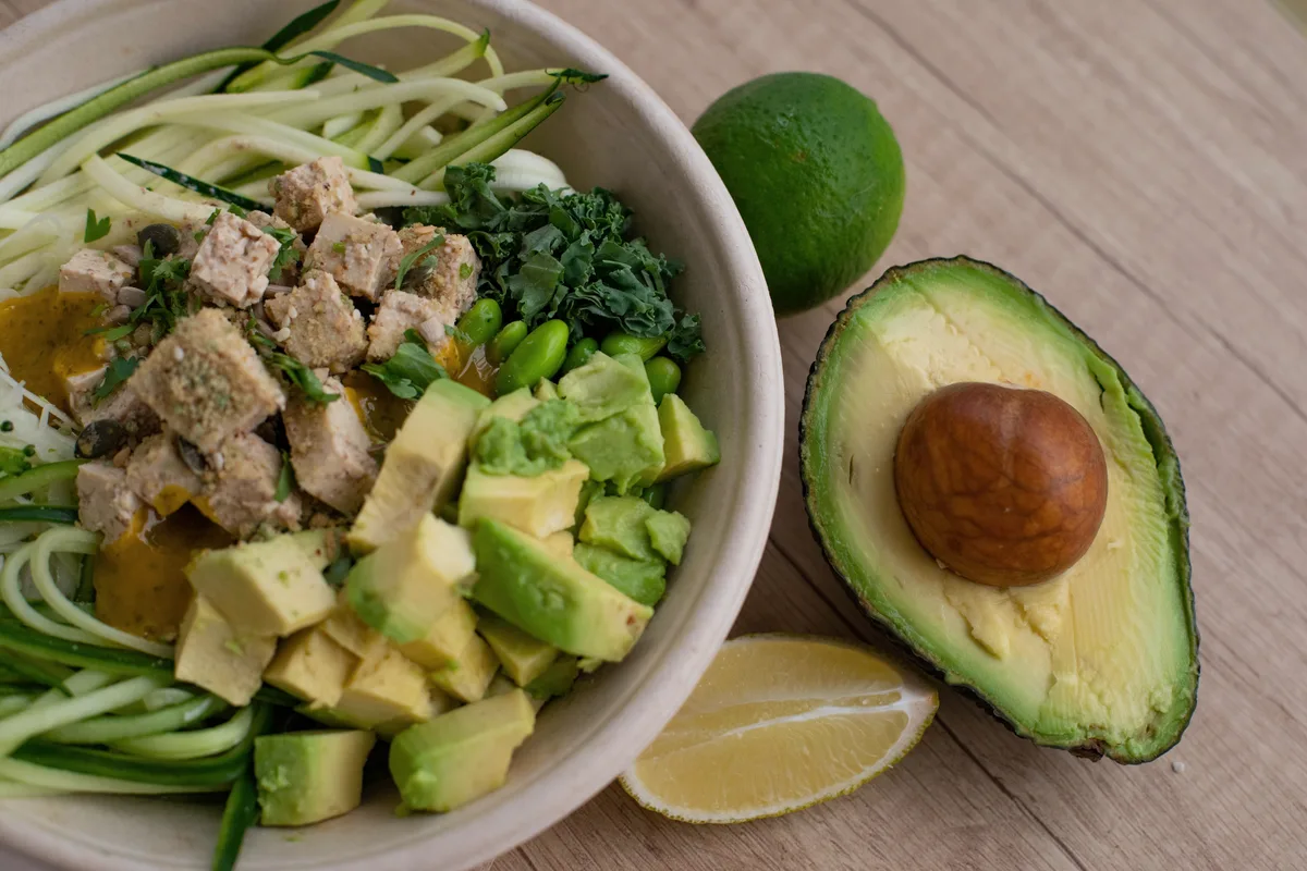 A healthy bowl of tofu, avocado, edamame and greens beside a halved avocado and lime